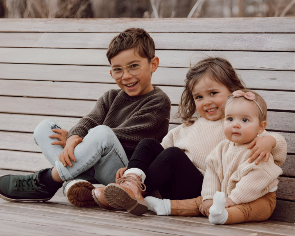 Three children sitting on a wooden bench outdoors in oh bebe knit sweaters