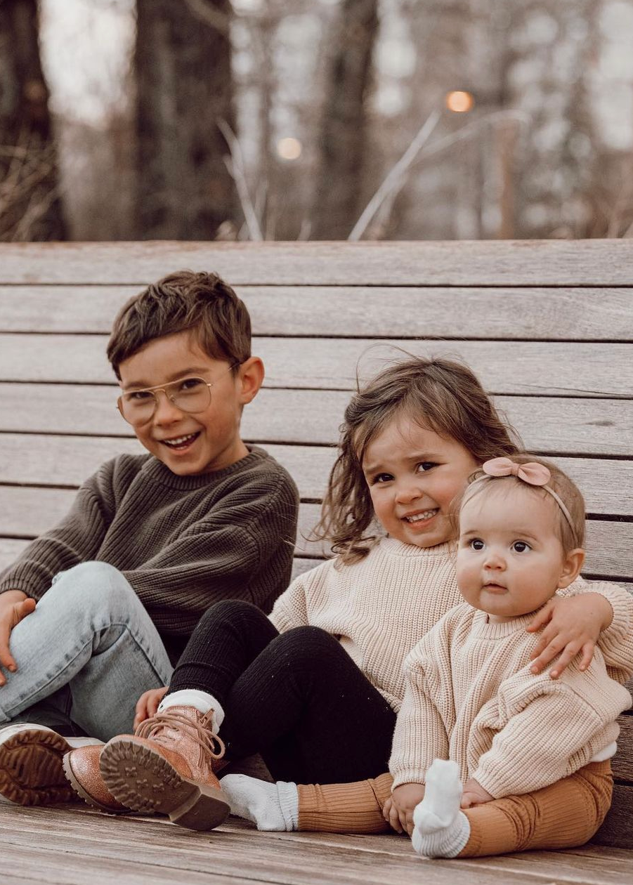 Three children sitting on a wooden bench outdoors wearing oh bebe sweaters